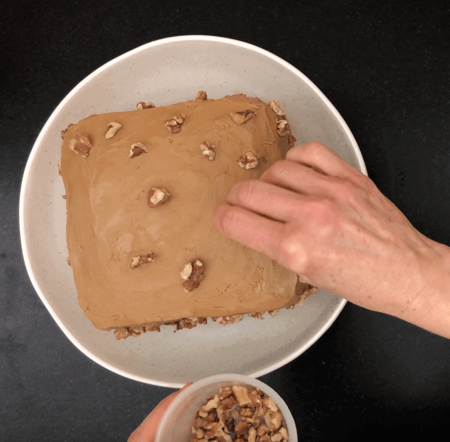 Square cake with coffee icing being decorated with chopped wlanuts
