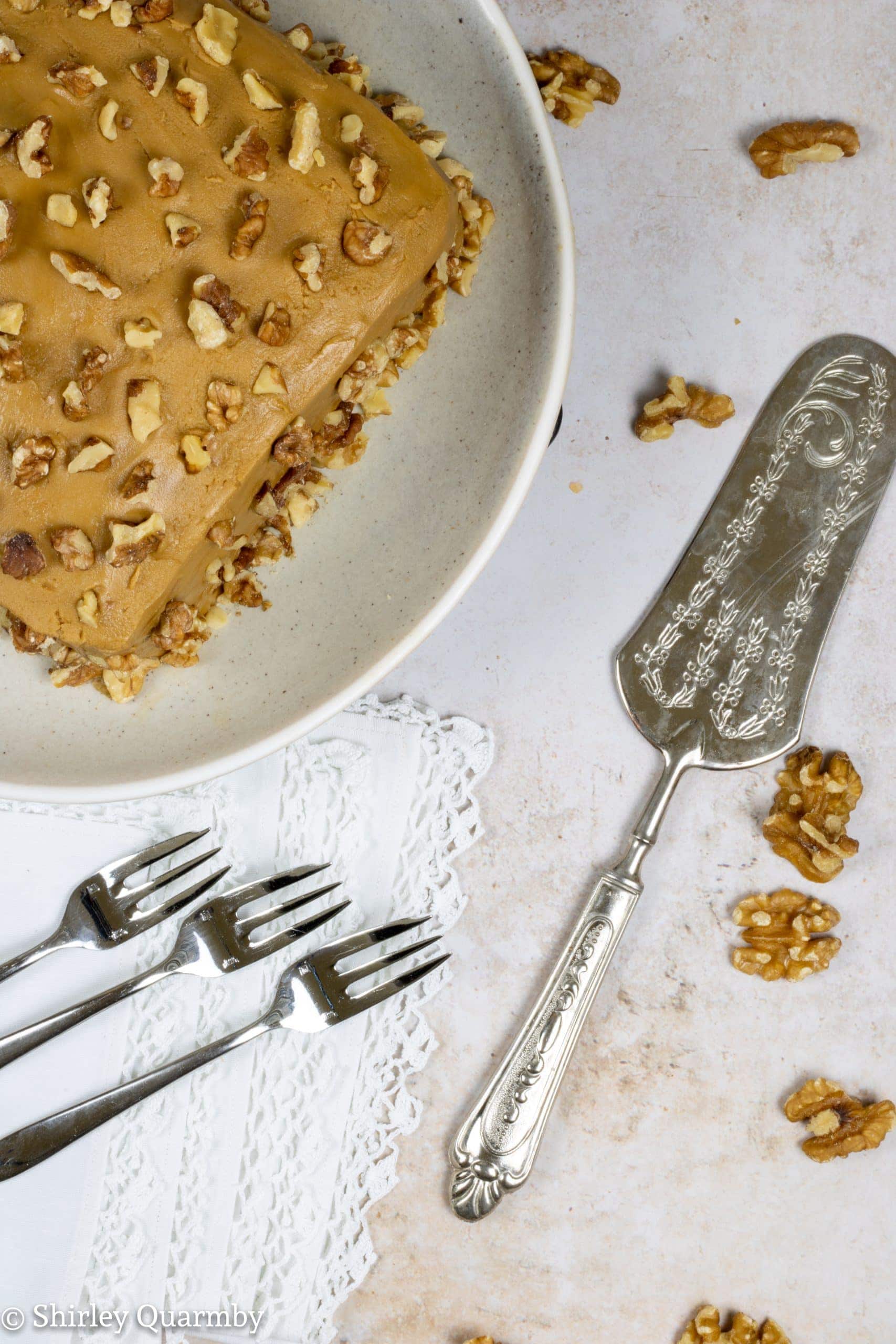 Square cake with golden brown icing decorated with walnut piece next to white napkins, serving forks and serving knife